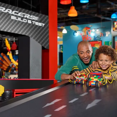 A dad and his son are building a LEGO race car together at LEGOLAND Discovery Center Bay Area. The dad is helping his son assemble the car using colorful LEGO bricks, while both focus on the creative process. The scene is set within a bright, interactive area of the center, with other LEGO structures and activities visible in the background. The father and son share a moment of excitement and teamwork as they work on their race car creation.