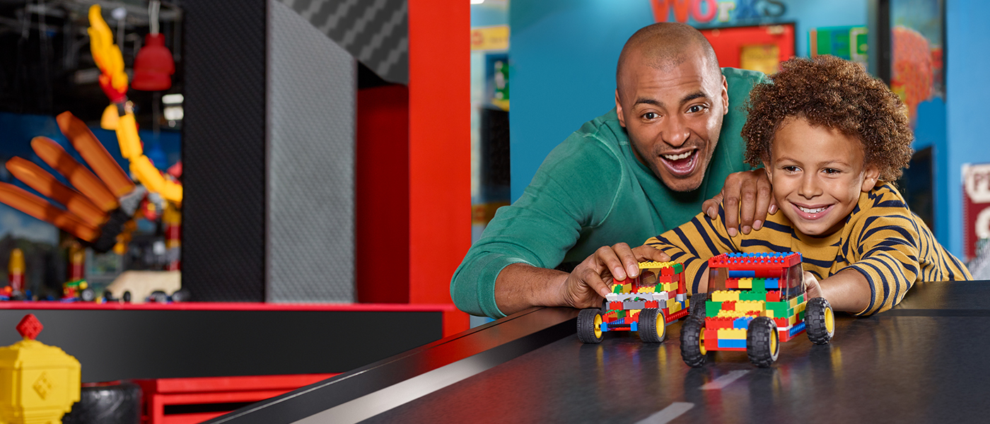 An excited adult and child racing colorful LEGO cars at the LEGOLAND Discovery Center's creative workshop.